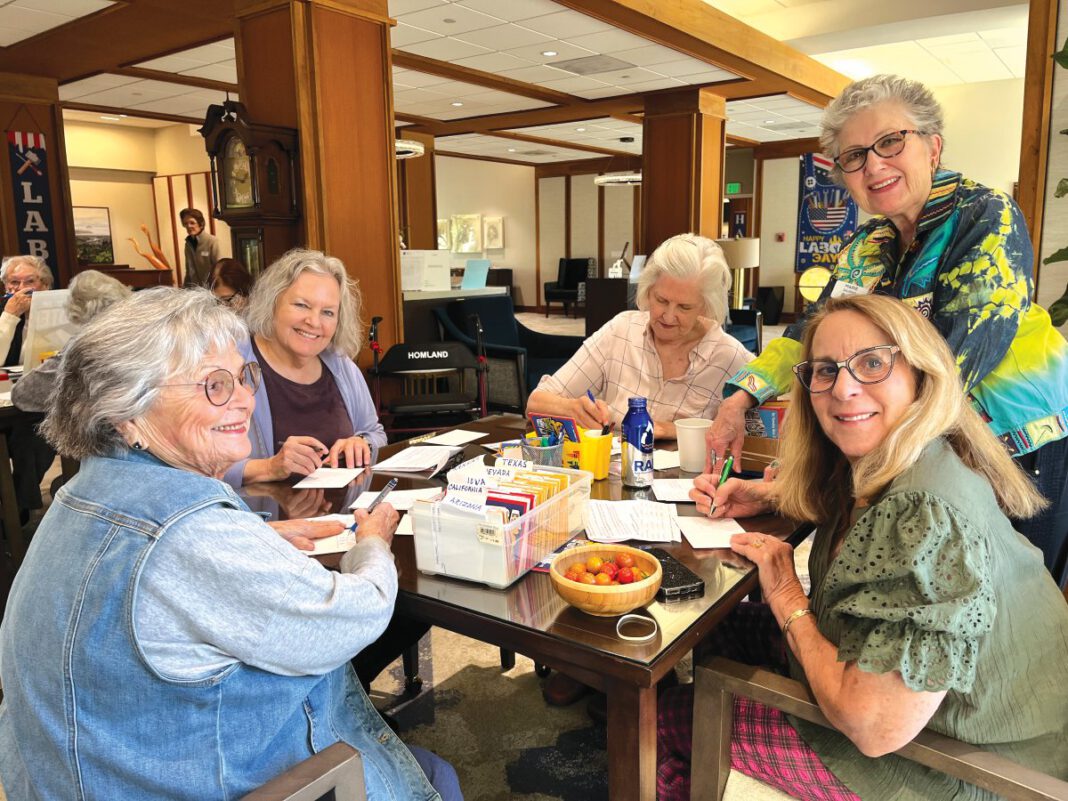 One woman standing with a group of women seated around a table writing postcards.