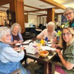 One woman standing with a group of women seated around a table writing postcards.
