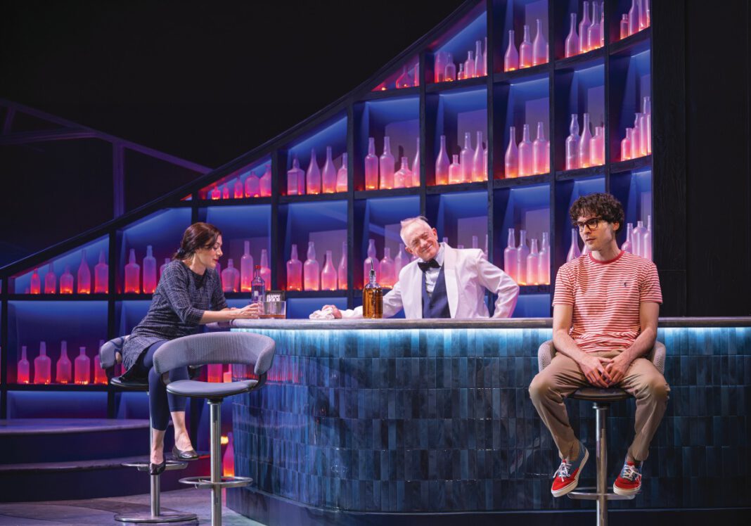 Three actors in a bar scene onstage, two seated on stools and one standing behind the bar, lit in blue and purple against shelves of glowing bottles.