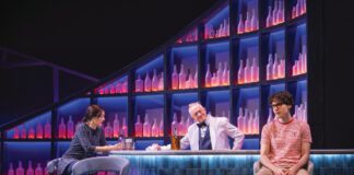 Three actors in a bar scene onstage, two seated on stools and one standing behind the bar, lit in blue and purple against shelves of glowing bottles.