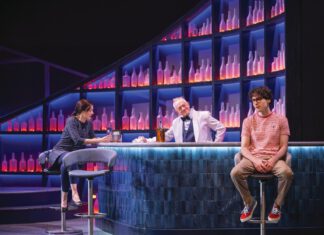 First Step: Dark Comedy Runs at Marin Theatre Three actors in a bar scene onstage, two seated on stools and one standing behind the bar, lit in blue and purple against shelves of glowing bottles.