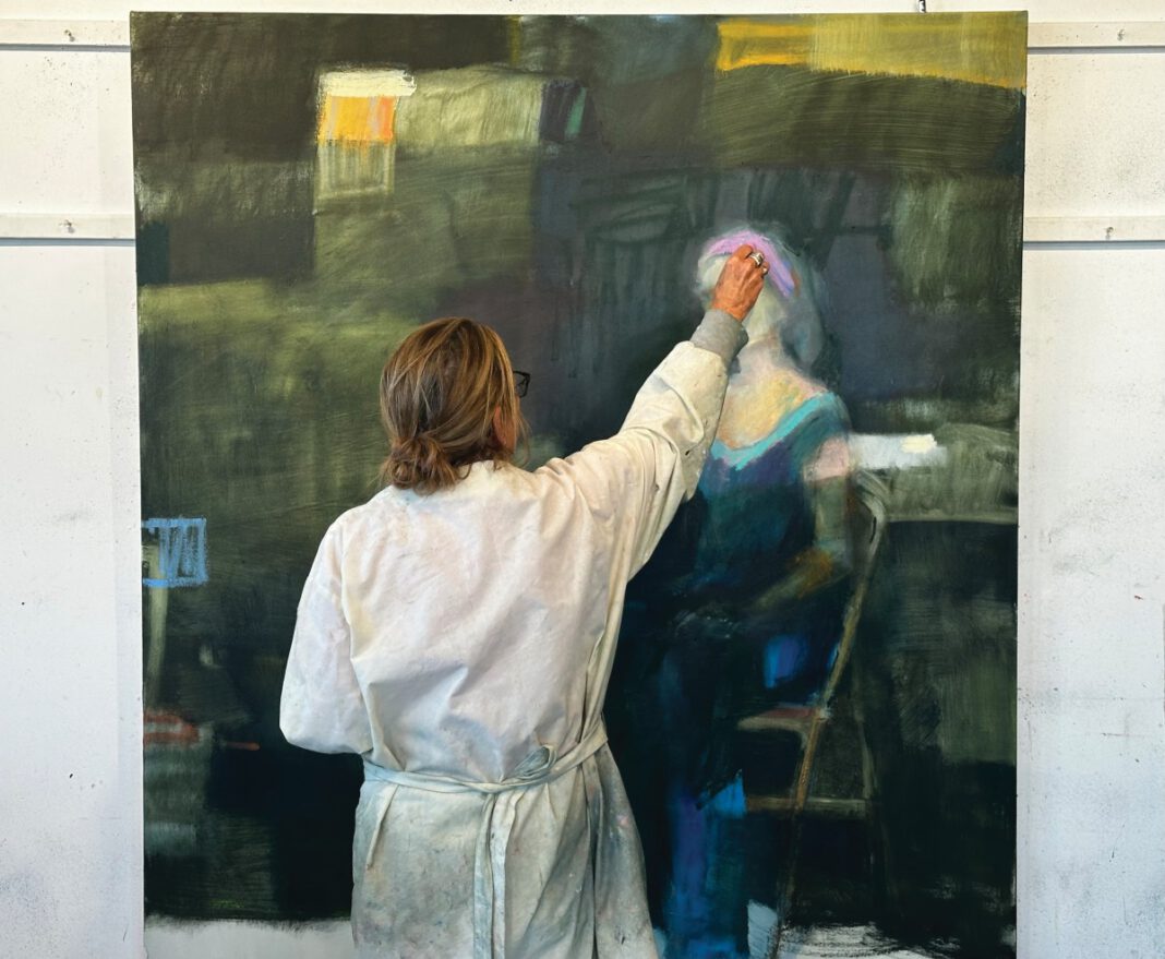 An artist in a paint-splattered smock reaches up to add paint to a large abstract portrait on canvas.