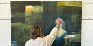 An artist in a paint-splattered smock reaches up to add paint to a large abstract portrait on canvas.
