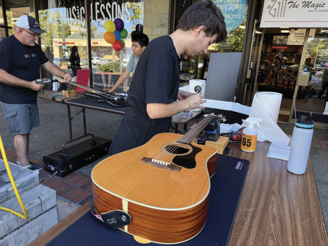 Two men restring guitars at an outdoor table in front of The Magic Flute music store.