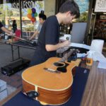 Two men restring guitars at an outdoor table in front of The Magic Flute music store.