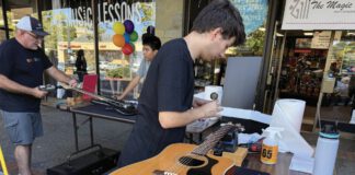 Two men restring guitars at an outdoor table in front of The Magic Flute music store.