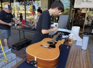 Two men restring guitars at an outdoor table in front of The Magic Flute music store.