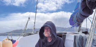 An older man in a ball cap sits in the cockpit of a sailboat, looking out over open water.