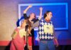 Three actors in costume perform on stage, two with raised arms and cardboard contestant numbers pinned to their chests.
