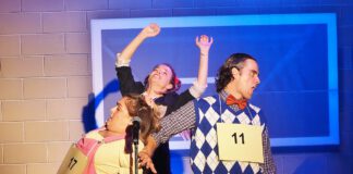 Three actors in costume perform on stage, two with raised arms and cardboard contestant numbers pinned to their chests.