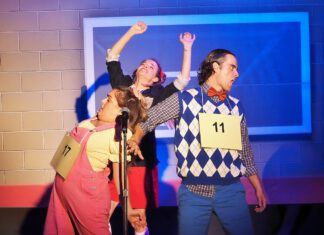 Three actors in costume perform on stage, two with raised arms and cardboard contestant numbers pinned to their chests.