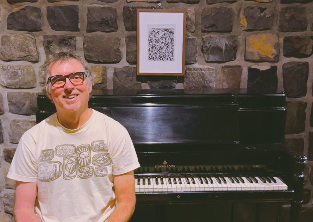 A smiling man in glasses and a graphic T-shirt stands in front of a Steinway upright piano against a stone wall.