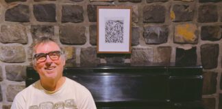 A smiling man in glasses and a graphic T-shirt stands in front of a Steinway upright piano against a stone wall.
