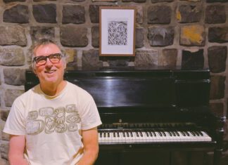 A smiling man in glasses and a graphic T-shirt stands in front of a Steinway upright piano against a stone wall.
