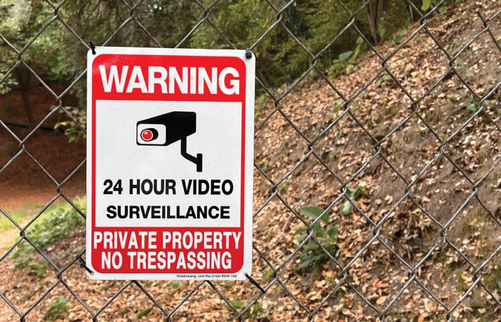 A red and white sign reading "24 Hour Video Surveillance, Private Property, No Trespassing" is attached to a chain-link fence.