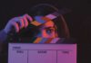 A person's eyes peek over a film clapperboard, one hand holding the slate open.