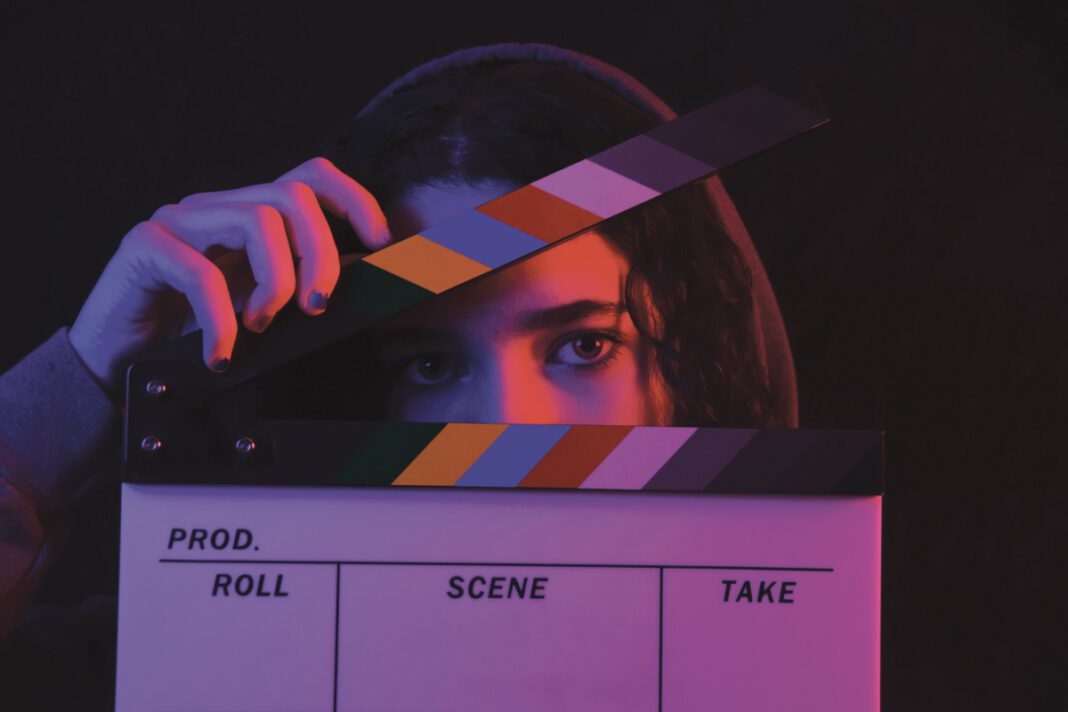 A person's eyes peek over a film clapperboard, one hand holding the slate open.