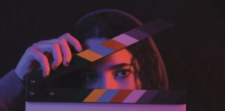 A person's eyes peek over a film clapperboard, one hand holding the slate open.
