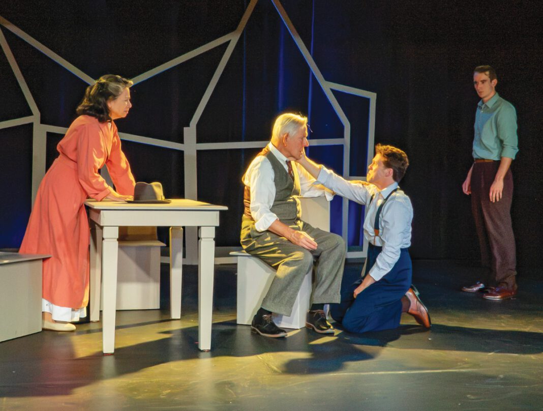 Four actors perform a on stage, with a young man kneeling to comfort an older seated man in a vest.