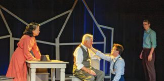 Four actors perform a on stage, with a young man kneeling to comfort an older seated man in a vest.