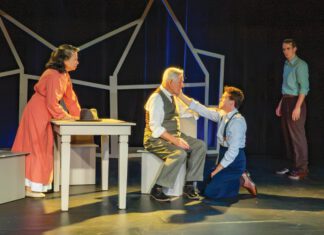 Four actors perform a on stage, with a young man kneeling to comfort an older seated man in a vest.