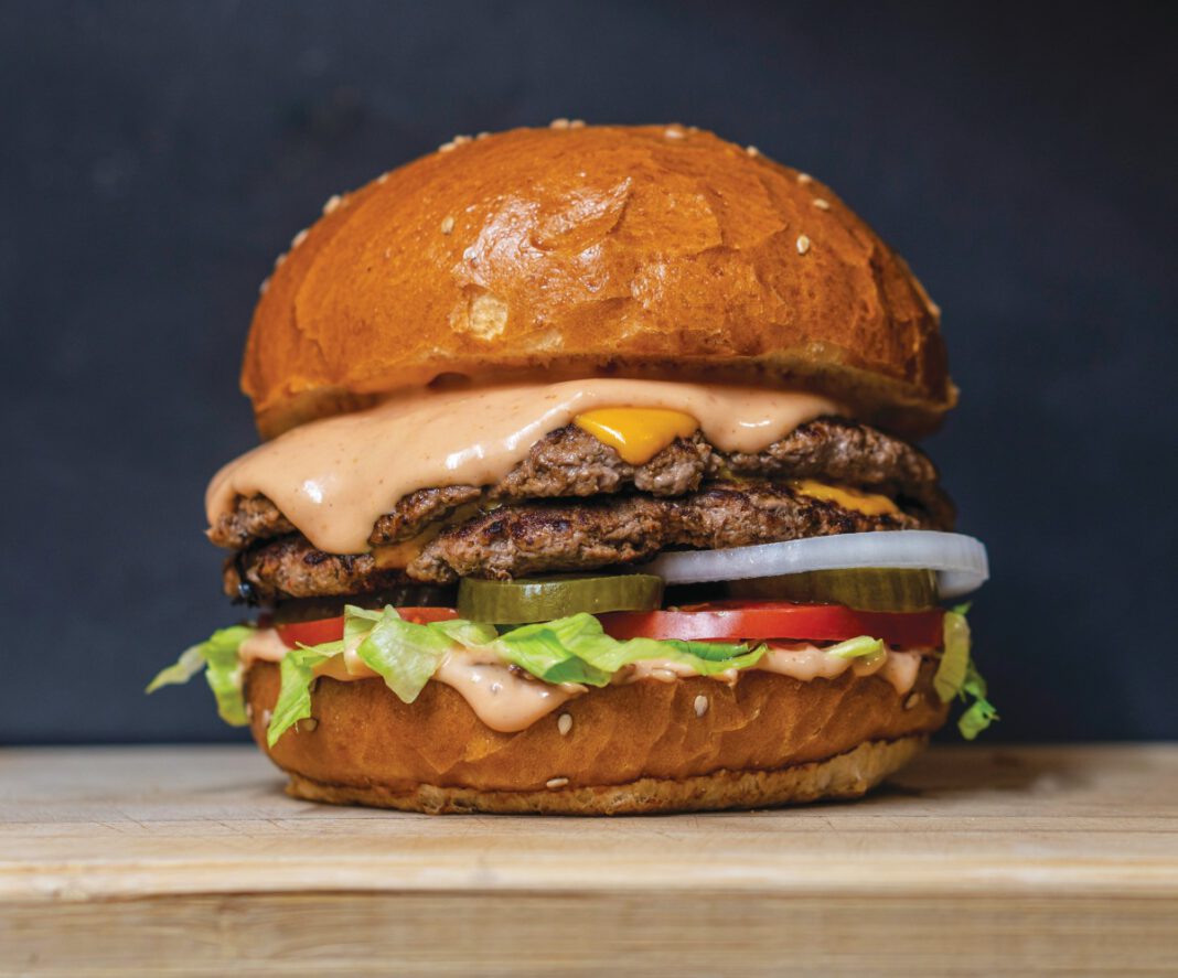 A double-patty cheeseburger with melted cheese, pink sauce, lettuce, tomato, pickles and onion on a sesame bun.