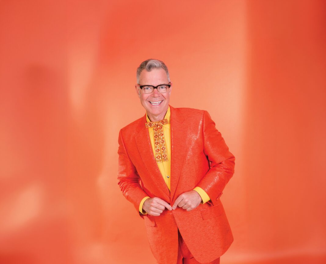 A smiling man with gray hair and black-framed glasses wears an orange blazer, yellow shirt and fancy orange and yellow tie, while standing in front of an orange background.