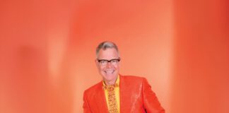 A smiling man with gray hair and black-framed glasses wears an orange blazer, yellow shirt and fancy orange and yellow tie, while standing in front of an orange background.