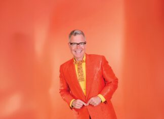A smiling man with gray hair and black-framed glasses wears an orange blazer, yellow shirt and fancy orange and yellow tie, while standing in front of an orange background.