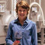 A smiling woman in a denim shirt holds a glass of red wine in front of large stainless steel tanks.