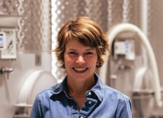 A smiling woman in a denim shirt holds a glass of red wine in front of large stainless steel tanks.