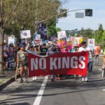 No Kings, Flock Cameras and Family Trees People marching on a street carry a red banner reading “NO KINGS” and others behind them hold protest signs.