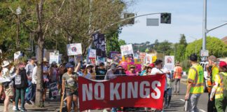 People marching on a street carry a red banner reading “NO KINGS” and others behind them hold protest signs.