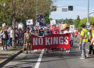 No Kings, Flock Cameras and Family Trees People marching on a street carry a red banner reading “NO KINGS” and others behind them hold protest signs.