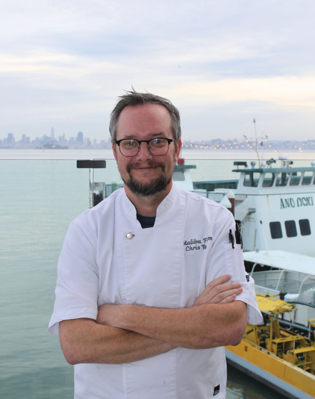 A smiling man with graying hair, eyeglasses and a beard stands with his arms crossed wearing a white chef’s coat.