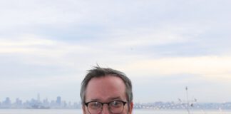 A smiling man with graying hair, eyeglasses and a beard stands with his arms crossed wearing a white chef’s coat.