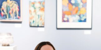 A smiling woman with long brown hair, eyeglasses and a nose ring sits on a wooden bench, holding two catalogs titled “Art at the Source: Open Studios.”