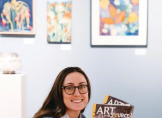 Linked Hands: Penelope Lenaerts of Art Trails Open Studios A smiling woman with long brown hair, eyeglasses and a nose ring sits on a wooden bench, holding two catalogs titled “Art at the Source: Open Studios.”
