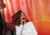 A woman with curly auburn hair wears a white dress and sings into a microphone, keeping her eyes closed.