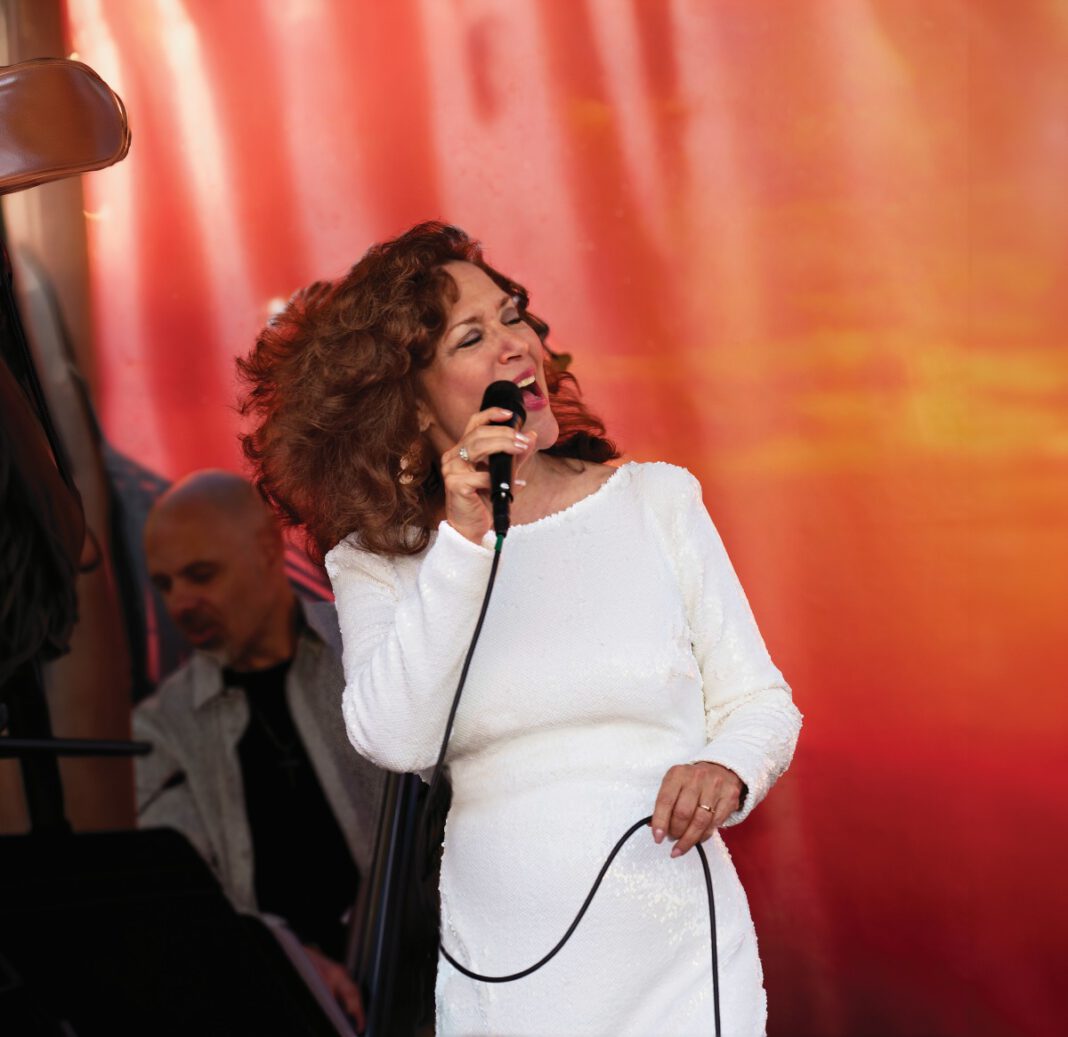 A woman with curly auburn hair wears a white dress and sings into a microphone, keeping her eyes closed.