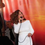 Divine Divas, Spanglish and Haunted Wine A woman with curly auburn hair wears a white dress and sings into a microphone, keeping her eyes closed.