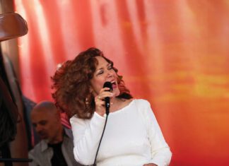 Divine Divas, Spanglish and Haunted Wine A woman with curly auburn hair wears a white dress and sings into a microphone, keeping her eyes closed.