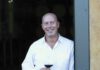 A smiling man dressed in a white shirt holds a glass of red wine as he leans against an open door.
