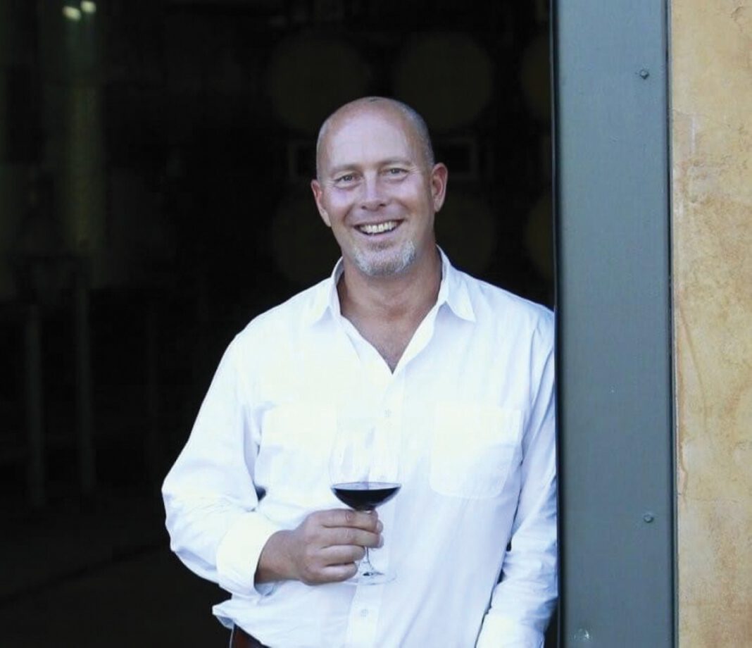 A smiling man dressed in a white shirt holds a glass of red wine as he leans against an open door.