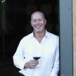 Full Circle, James MacPhail Looks Back, Ahead A smiling man dressed in a white shirt holds a glass of red wine as he leans against an open door.