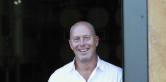 A smiling man dressed in a white shirt holds a glass of red wine as he leans against an open door.