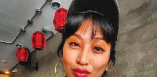 A woman with black bangs is wearing a black cap, an orange shirt, a black vest and gold hoop earrings and she sports orange eyeshadow, red lipstick and arm tattoos.