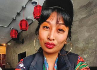 A woman with black bangs is wearing a black cap, an orange shirt, a black vest and gold hoop earrings and she sports orange eyeshadow, red lipstick and arm tattoos.