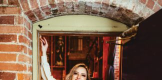 A smiling woman with long blonde hair, a white shirt and black leather pants leans against a brick wall.