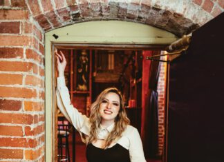 Sound Decisions: Announcing Our 2026 North Bay Music Awards Winners A smiling woman with long blonde hair, a white shirt and black leather pants leans against a brick wall.